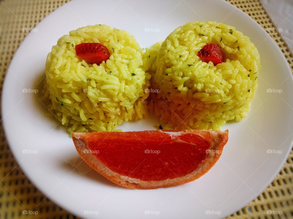 Yellow rice in the form of two eyes with slice of strawberries and one slice of grapefruit. In a white round plate background.