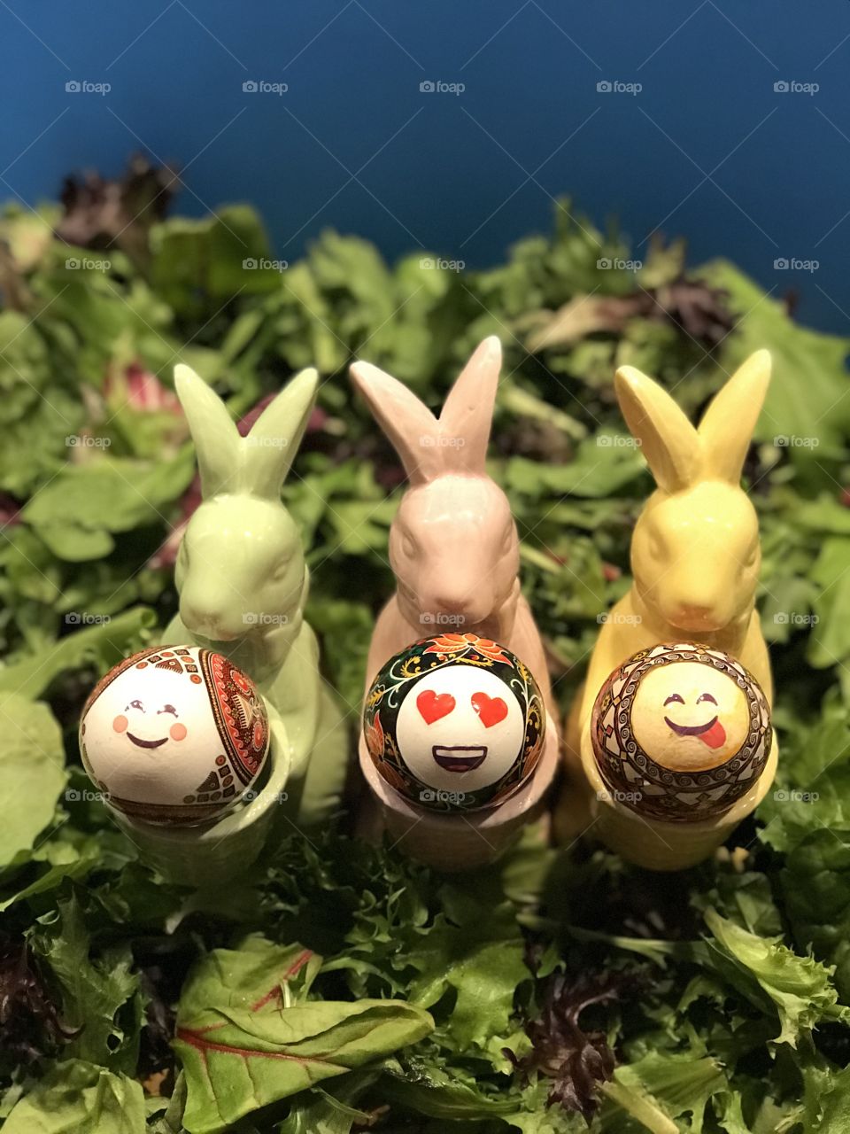 Three Easter bunny egg holders holding Ukrainian decorated eggs with faces all on a bed of greens. 