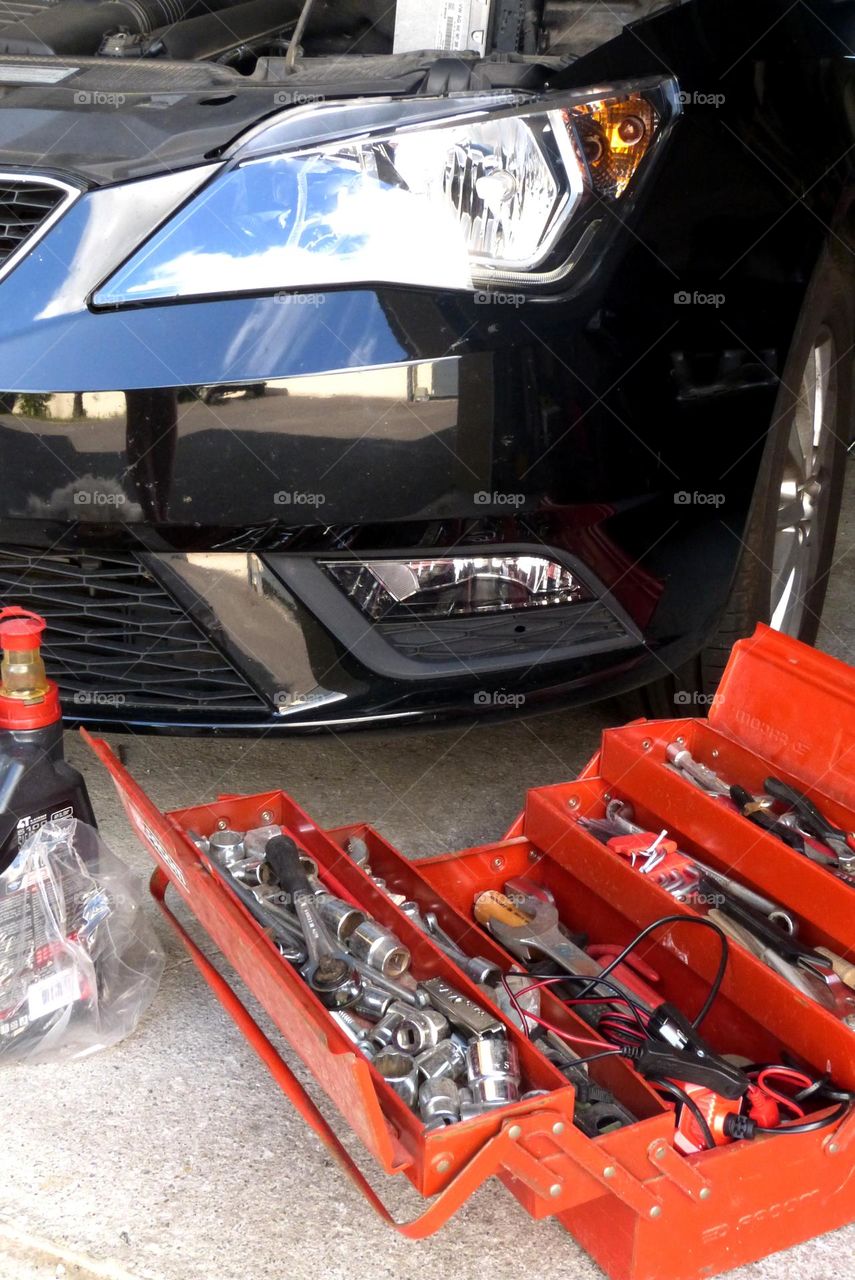 In a garage, a red-colored toolbox is open in front of a black car with the hood raised. the floor is cement.