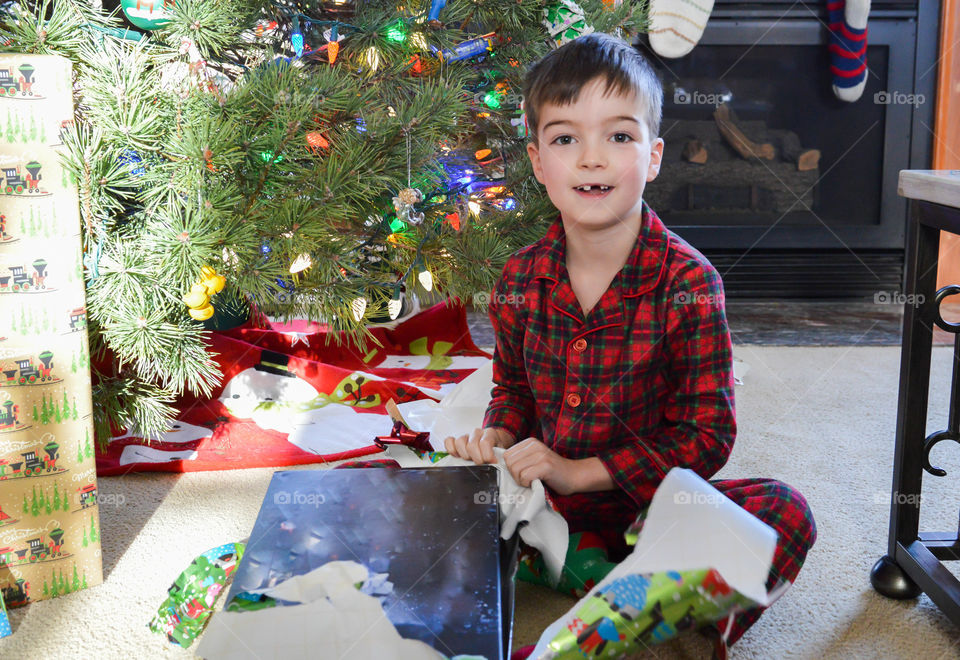 Young boy with a missing tooth smiling and opening presents on Christmas morning in front of a Christmas tree indoors