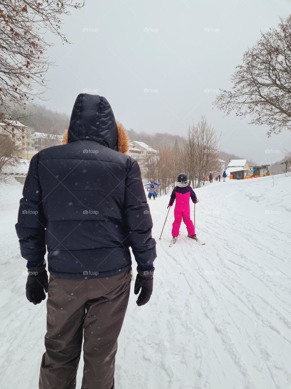 dad from behind is watching his little girl learning to ski in the snow