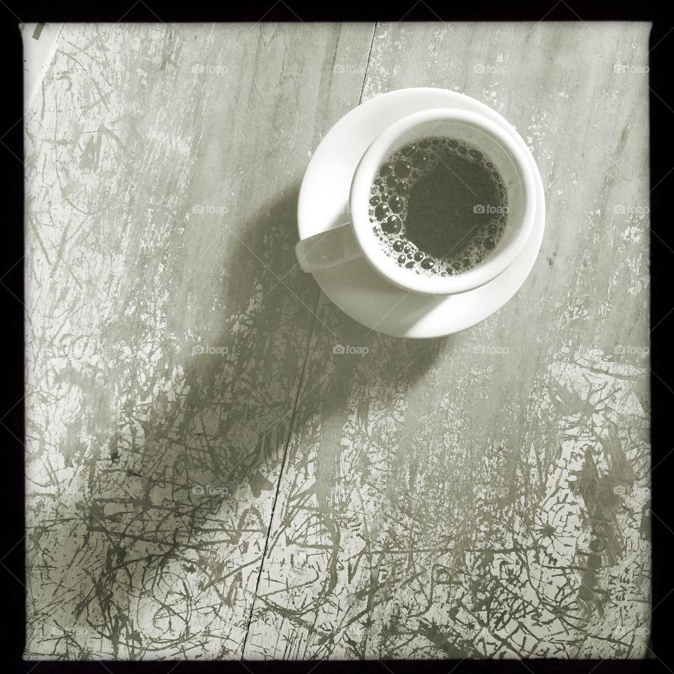 A simple cup of black coffee sits on a worn wooden table. A stream of morning sunlight casts a long, low shadow across the table.