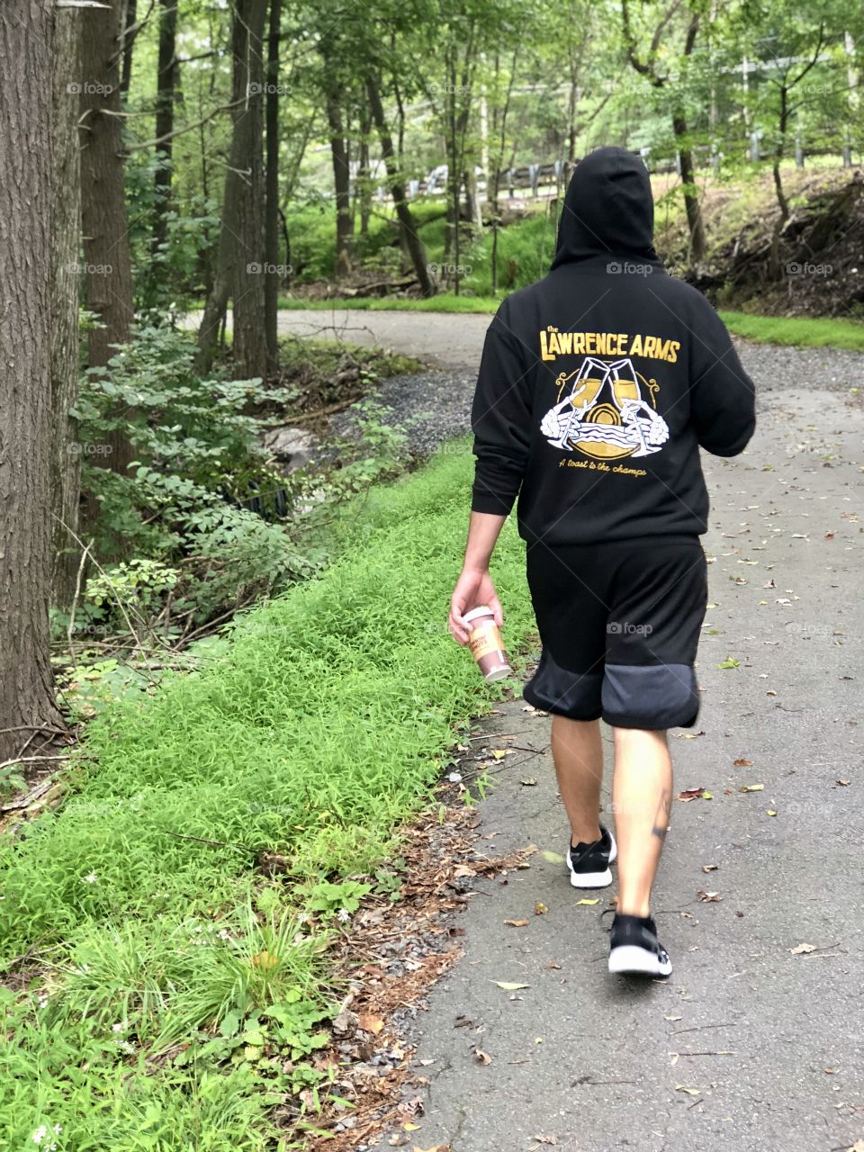 Young man leisurely strolling in front of me on a cold summer morning in shorts and a hoodie, drinking a coffee and texting on his phone. Daily exercise. 