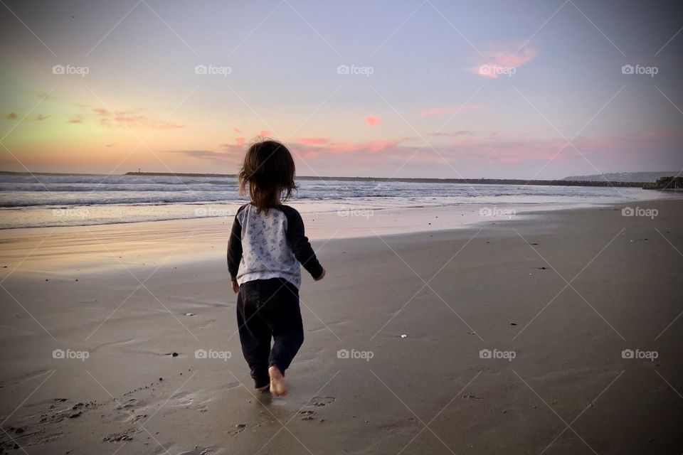 A young child walks along a California beach during sunset, soft waves illuminated in the background.