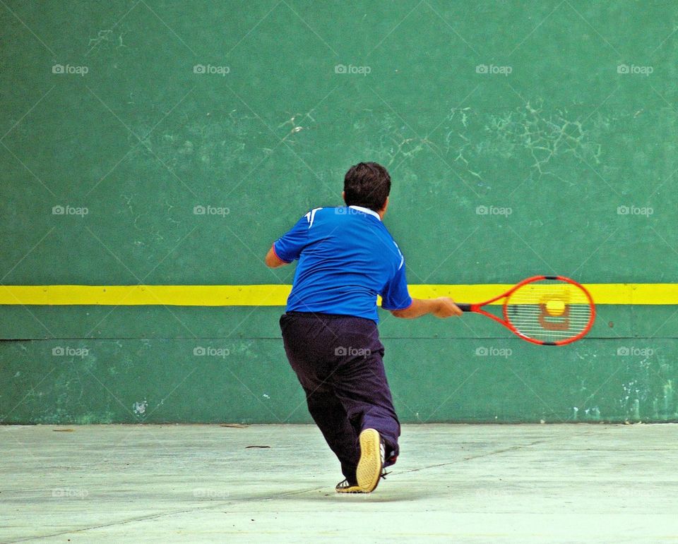 A tennis player viewed from the back swinging his racket and connecting with ball on the center of the racket.