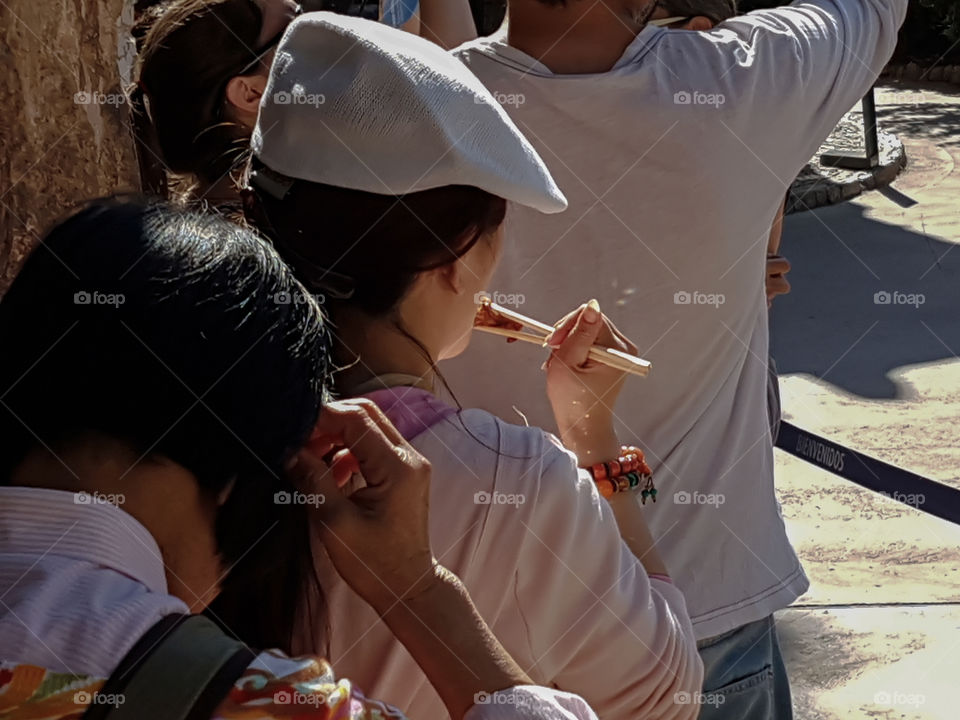 A young Asian female is eating with wooden chopsticks while waiting at an outdoors museum queue line at Casa del Guarda Park Guell in Barcelona, Spain.