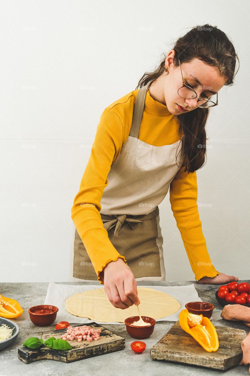 Beautiful caucasian teenager girl in a yellow sweater and apron stands at the table and cooks pizza on the white wall in the kitchen, side view close-up. Concept of cooking homemade pizza, favorite spot at home.