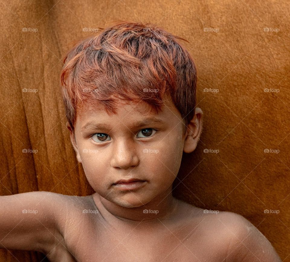 This close-up portrait features a young boy with striking reddish-brown hair, standing shirtless against a backdrop of earthy tones, likely the side of an animal such as a cow. His deep, expressive eyes stare directly at the camera,