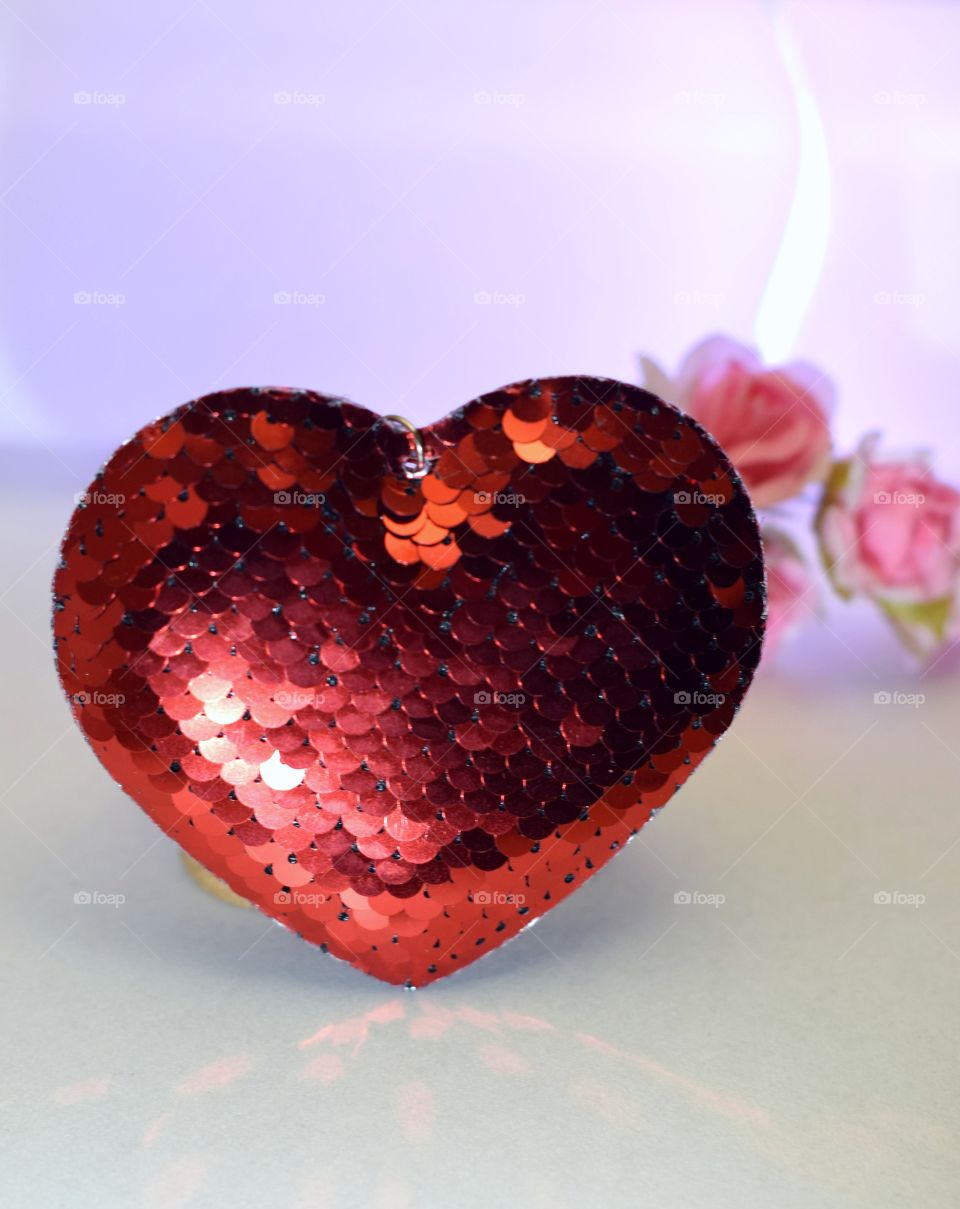 Red heart from sequins on a light background. In the background a bouquet of flowers in blur