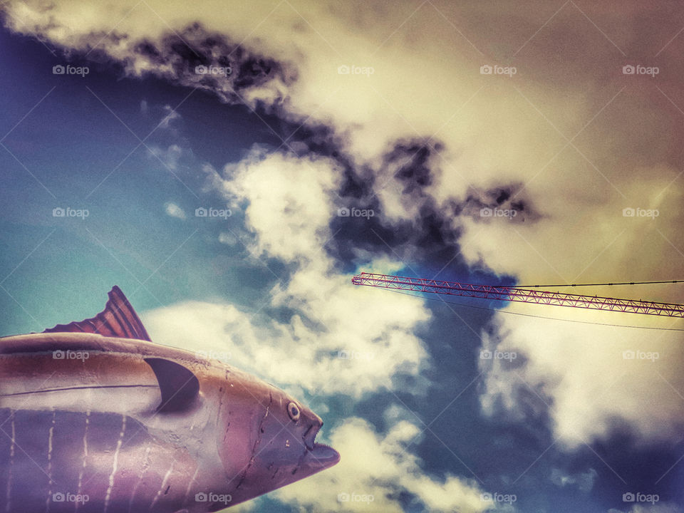 A fish sculpture and a construction crane against a cloudy sky