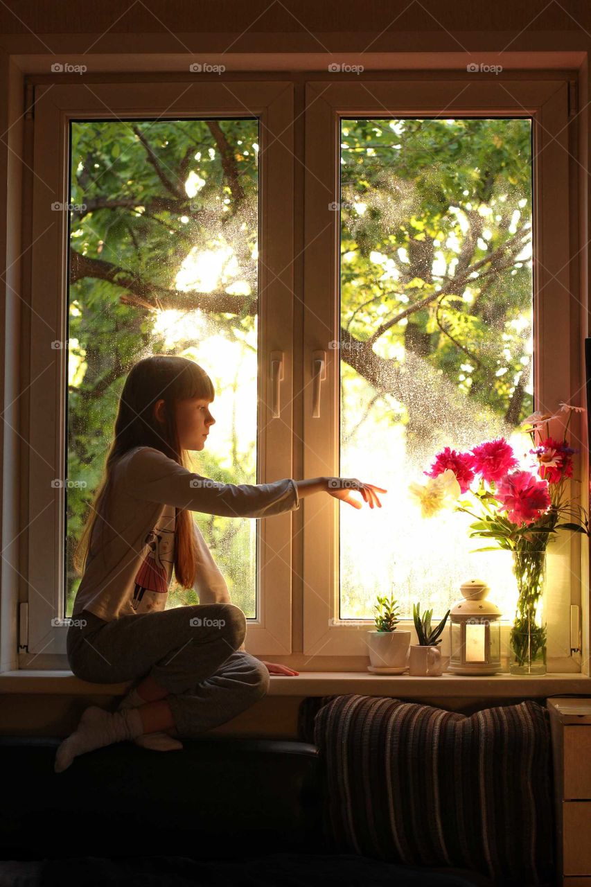 Little girl sits by the window and reaches for peonies