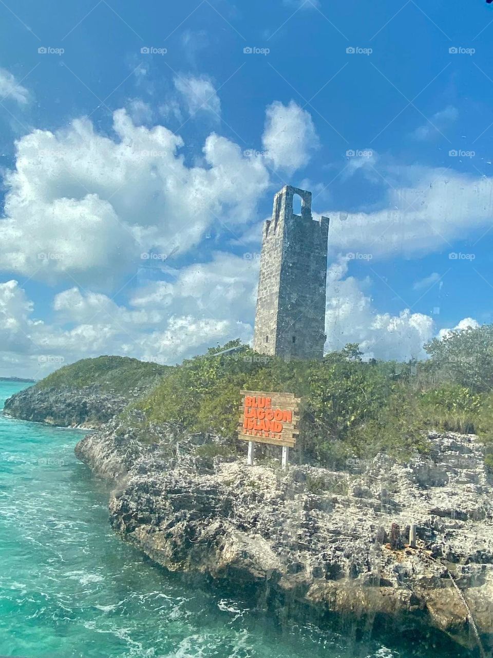 A rocky shore hosts a tower amidst the blue skies filled with fluffy white clouds. The water is a spectacular shade of aquamarine. 