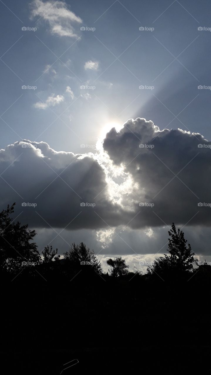 sun looking through heavy rain clouds illuminating blue sky with black silhouettes of trees