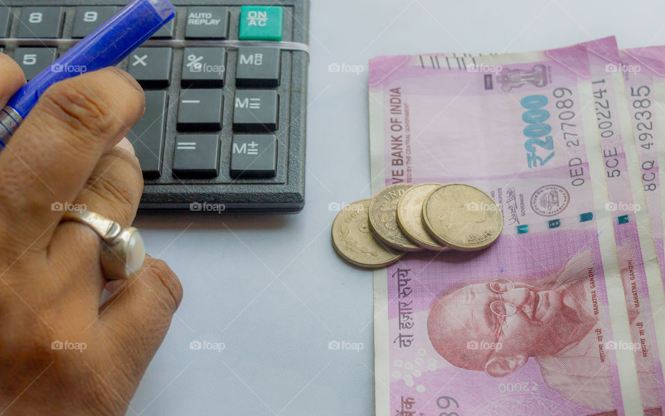 Cropped hand of a woman checking account with calculator and holding a pen. Indian currency notes and coins are on the side of table. Corporate business concept.
