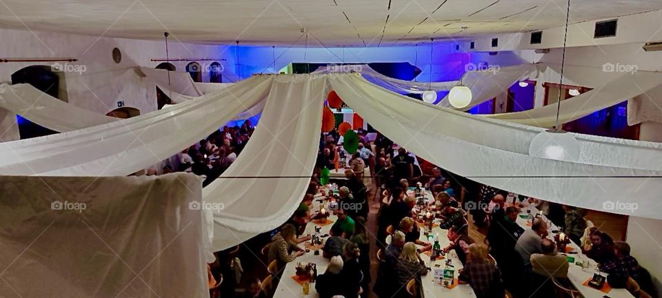 White fabric and festive girlands are draped across the hall on lines near the ceiling. This is to outfit the evening of “Irish Folklore” with song, dance and (not to forget) food in “Bavaria” with the proper decoration. 2023. Hypnotic Productions