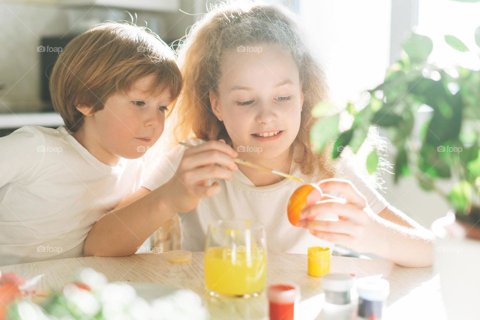 Two siblings brother and sister toddler boy tween girl painting easter eggs on kitchen at home on the spring sunny day