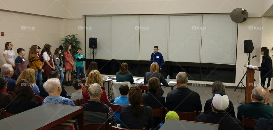 Middle public school age children at a spelling bee; public speaking event with a large crowd and neutral background.