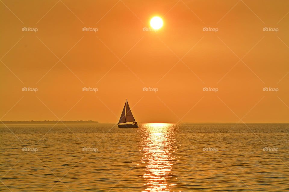 A beautiful sunset scene featuring a solitary sailboat on the water. The sun, low on the horizon, casts a warm, golden glow across the sky and reflects on the calm water, creating a serene and picturesque setting