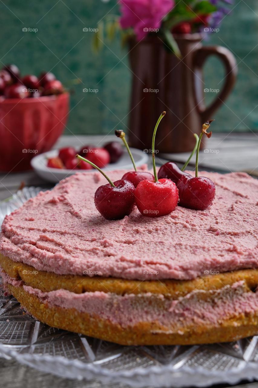 Four bright red cherries on top of an almond and cherry cake and in the background a red bowl with cherries is a clay jug with country flowers