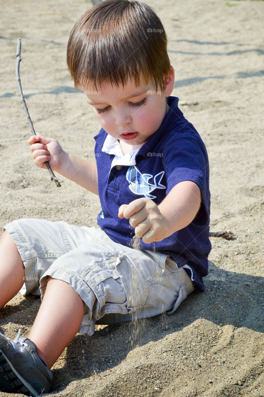 Toddler boy sitting and playing in a sand box while holding a stick