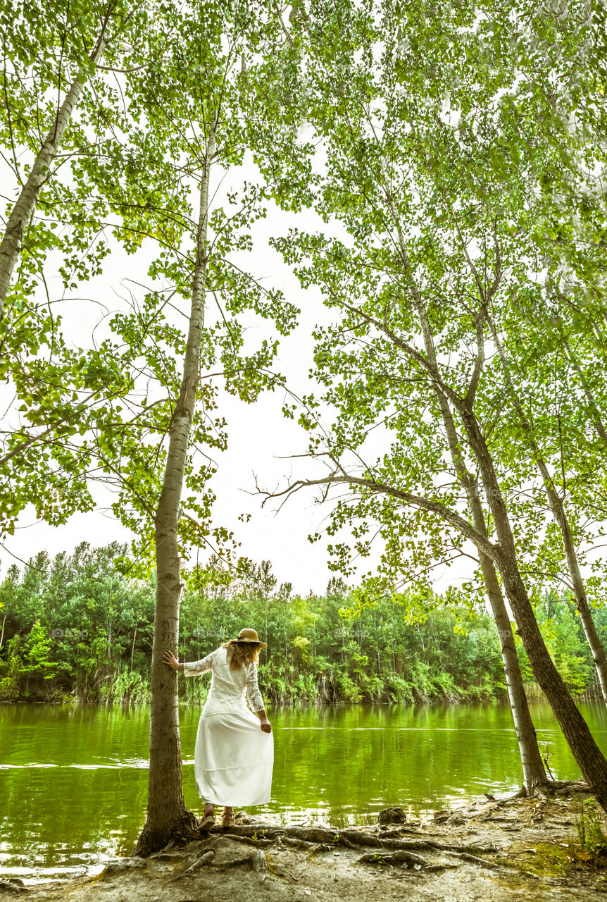 Woman from behind in white dress wearing straw hat, standing in front of the lake, in summer green forest landscape at afternoon, enjoying nature. Freedom, romance, resting and tranquility. Lonely female In thought, in the trees at the park.