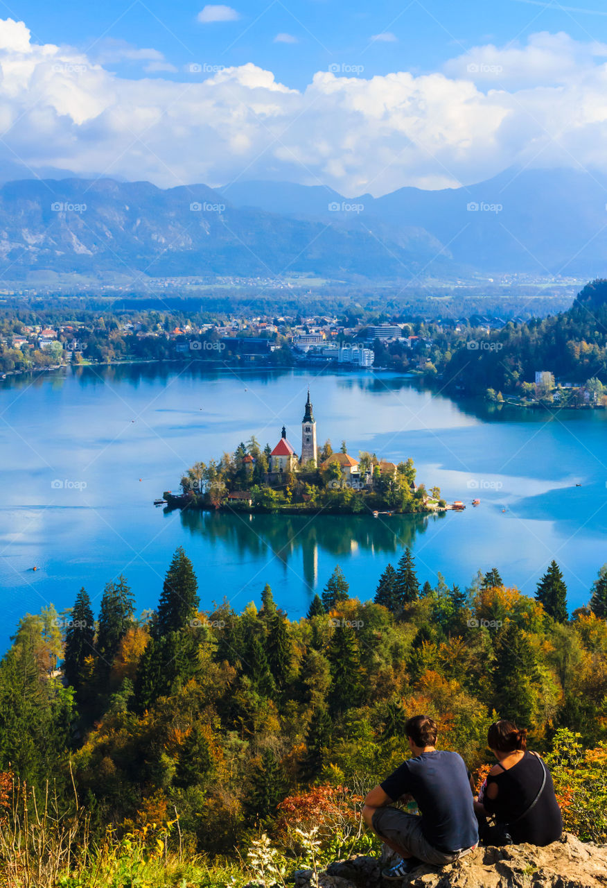 Romantic destination. Couple sitting on rock on top of hill, enjoying the view of lake Bled in autumn.