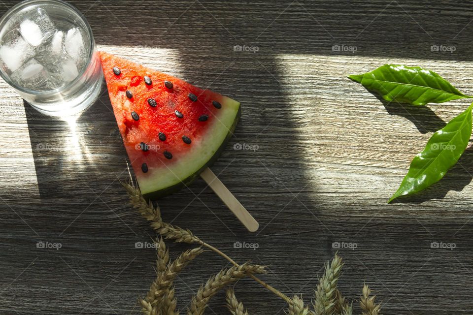 A piece of ripe, red watermelon and cold juice, a cocktail with ice and ears of wheat stand on a wooden table.  Summer drinks and snacks.