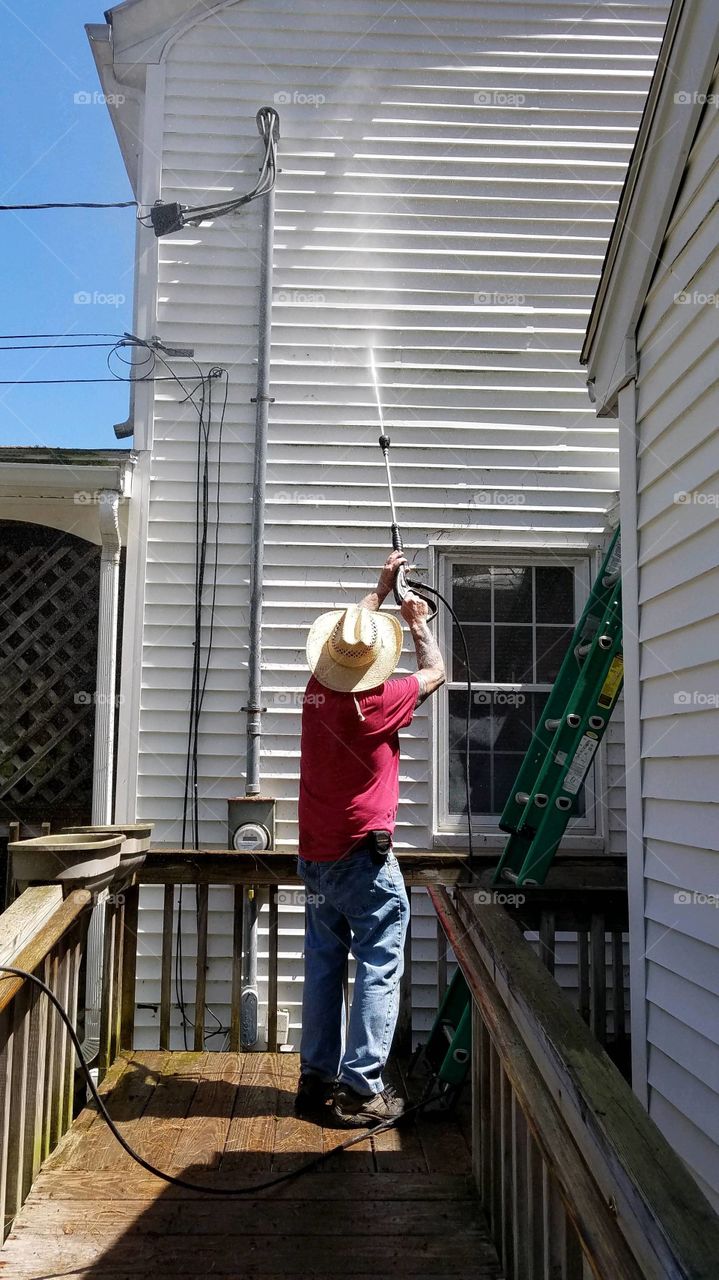 Man power washing house with vinyl siding, clean area seen making it easier to do.  Sunny warm day for doing this project.