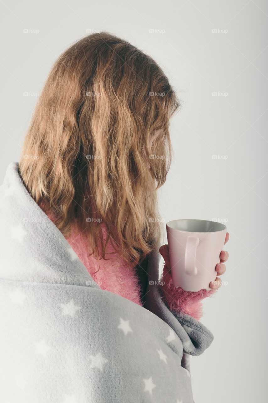 Young woman has caught cold, has day off, sick day, wrapped in blanket, holds cup of herbal tea. Profile portrait view