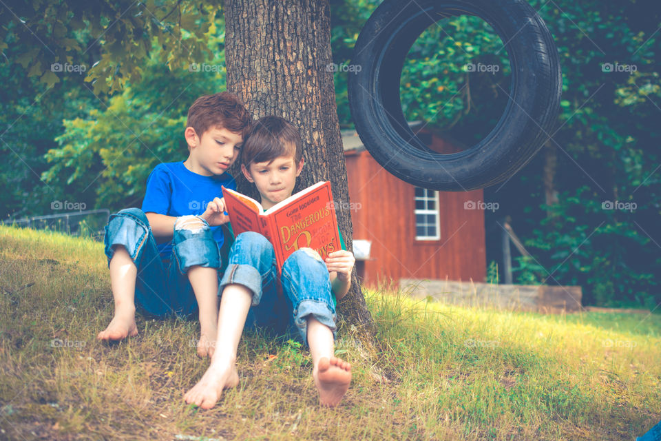 Two Young Boys Reading a Book Next to a Tree with Tire Swing