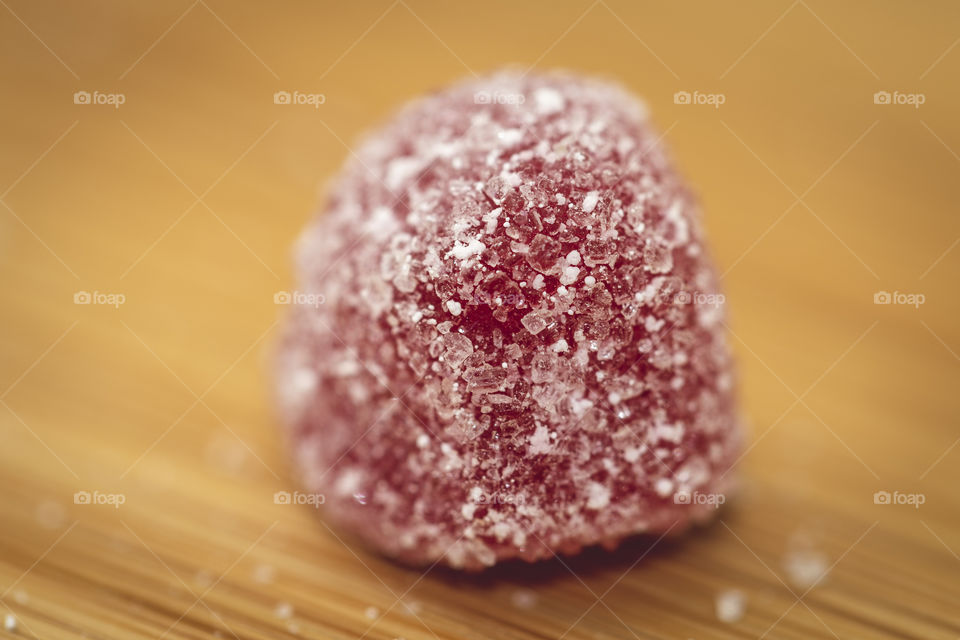 A macro portrait of a single red piece of sugar coated candy on a wooden table. It is a red tiny piece of candy with tiny bits of sugar on it.