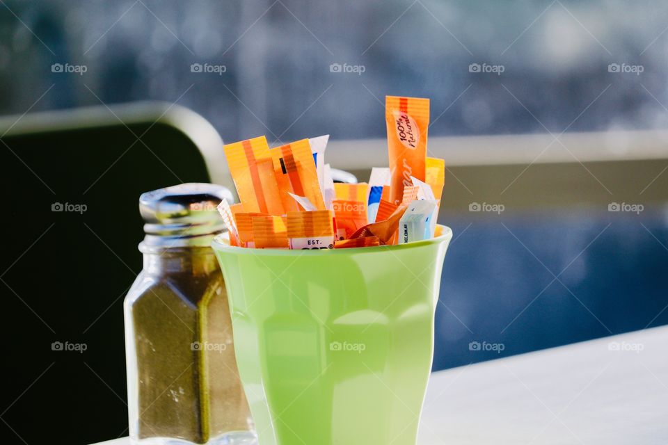 Condiments on restaurant table, glass stainless steel topped pepper shaker and lime green glass filled with selection of sugar in paper tubes, blurred background, copy and text space, hospitality and service industry 