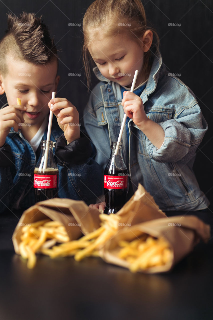 beautiful, stylish, fair-haired children, drinking Coca Cola, eating potatoes, dressed in denim clothes, free photo style.  boy and girl have fun, rejoice, beautiful advertisement for coca cola drink