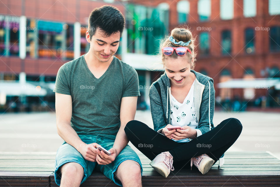 Couple of friends, teenage girl and boy, having fun together, using smartphones, sitting in center of town, spending time together. Real people, authentic situations