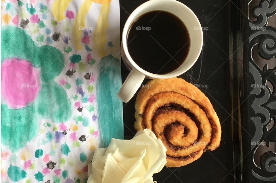 Breakfast for mom in bed. Black Coffee, cinnamon roll, white rose and child's painting on black tray. 