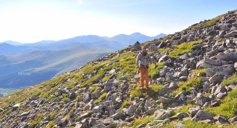 A man hikes with trekking poles through a mountain tundra in the high country on a very sunny day.
