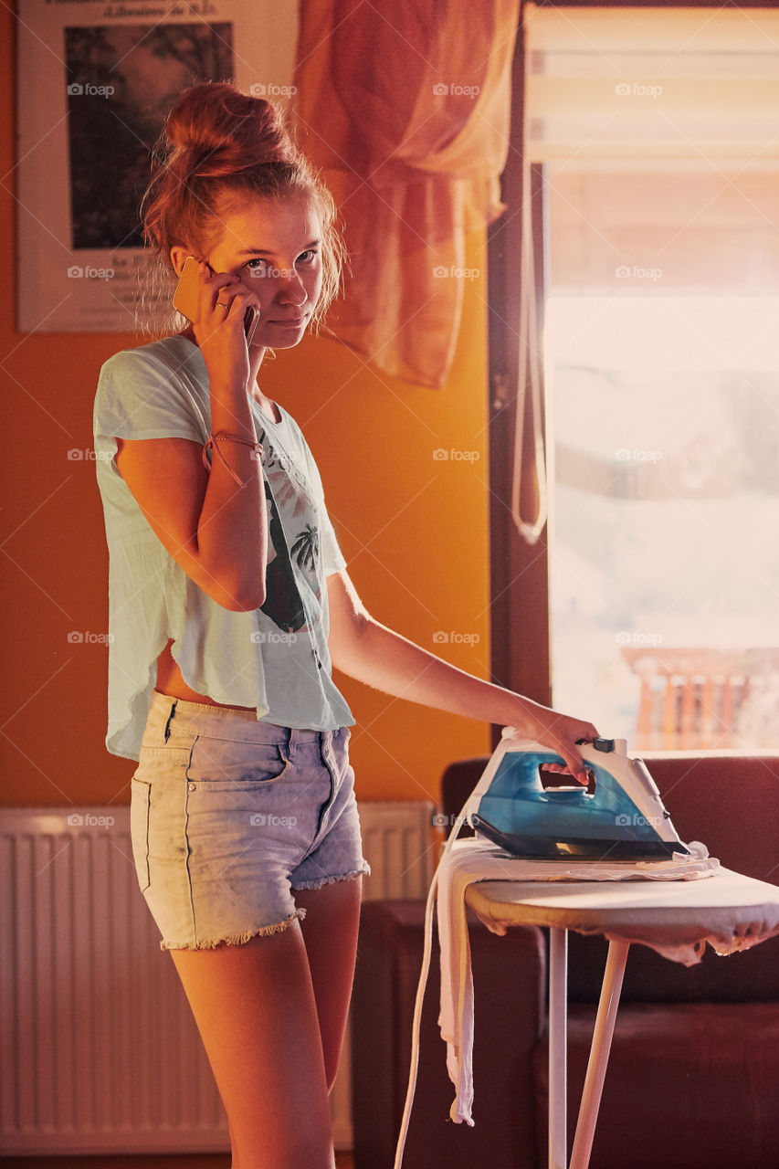 Young woman is ironing her clothes and talking on a smartphone simultaneously, standing by a ironing board in a room at home. Routine housekeeping task at home. Candid people, real moments, authentic situations
