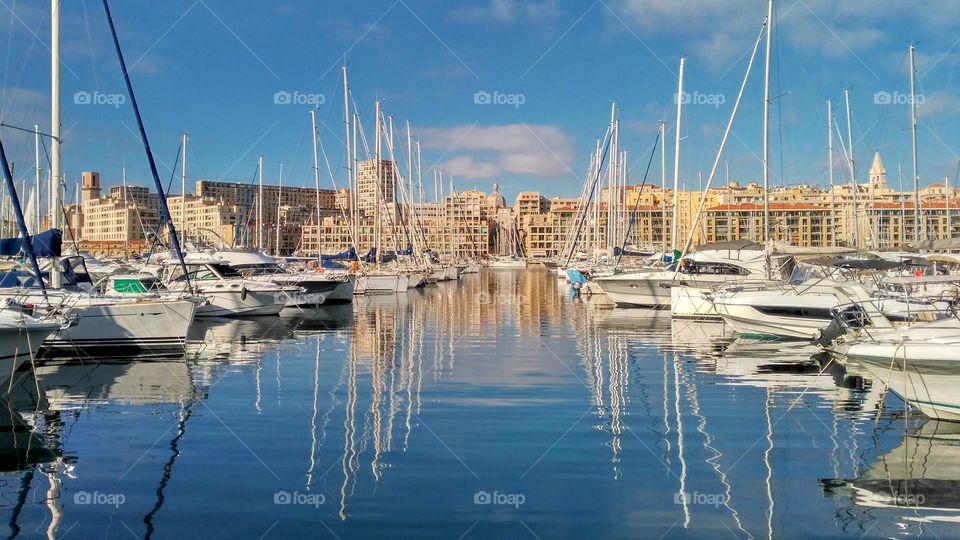 Old port of Marseille, France. In the background the buildings of the city, a blue sky with clouds and more closely boats with their silhouette reflected in the water like a fishbone.