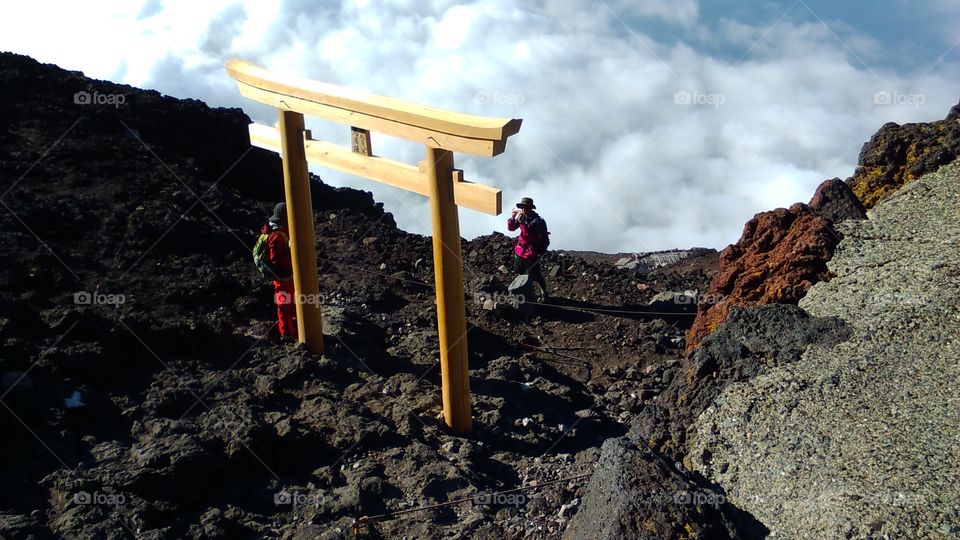 Above the late morning clouds, hikers pause for a photo with an elegant wooden gate near the top of Mount Fuji.