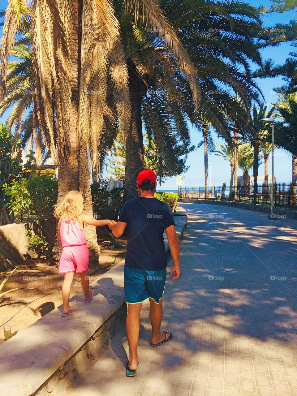 Man and blond little girl walking on a boardwalk with palm trees to the beach on a sunny day.
