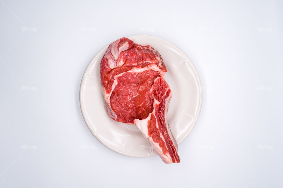 On a white background is a white plate on which lies a fresh, juicy piece of beef meat on a bone