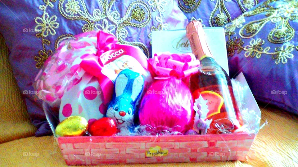 Easter basket with big and small Easter eggs, bubbly wine, Easter cake and chocolate Easter bunny surrounded by violet cushions