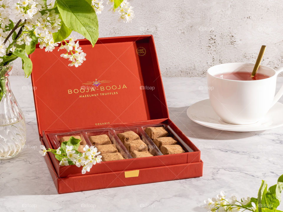 Chocolate truffles in a red box and a cup of tea on a white table, white spring flowers. 