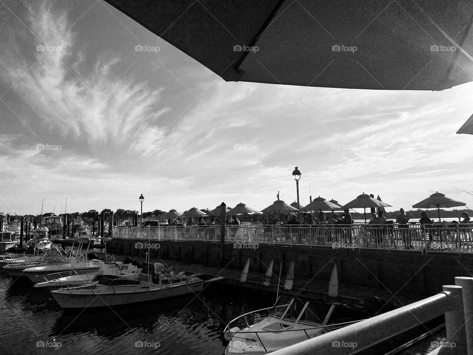 A black and white photo of boats docked in a marina taken at a bar/restaurant on the pier. Part of the umbrella that shades the table is overhead. This was a beautiful evening, and I think the sky is just stunning in this picture.