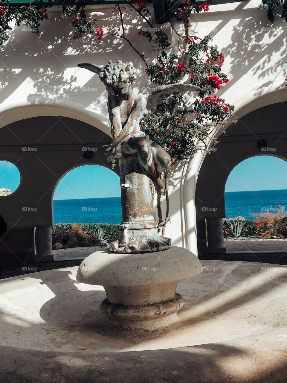 Shadows of leaves and wooden slats printed on the wall , an angel statue sitting on a fountain , behind is the blue sea 