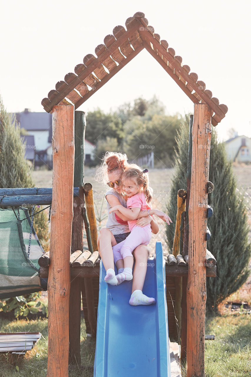 Teenage girl playing with her younger sister in a home playground in a backyard. Happy smiling sisters having fun on a slide together on summer day. Real people, authentic situations