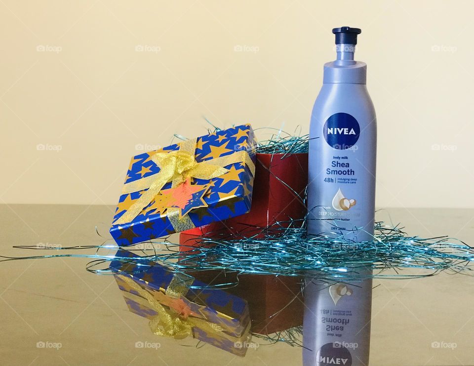 Nivea shea smooth body lotion and a blue gift box filled with blue glitter strips.and the Nivea lotion reflects on glass table.