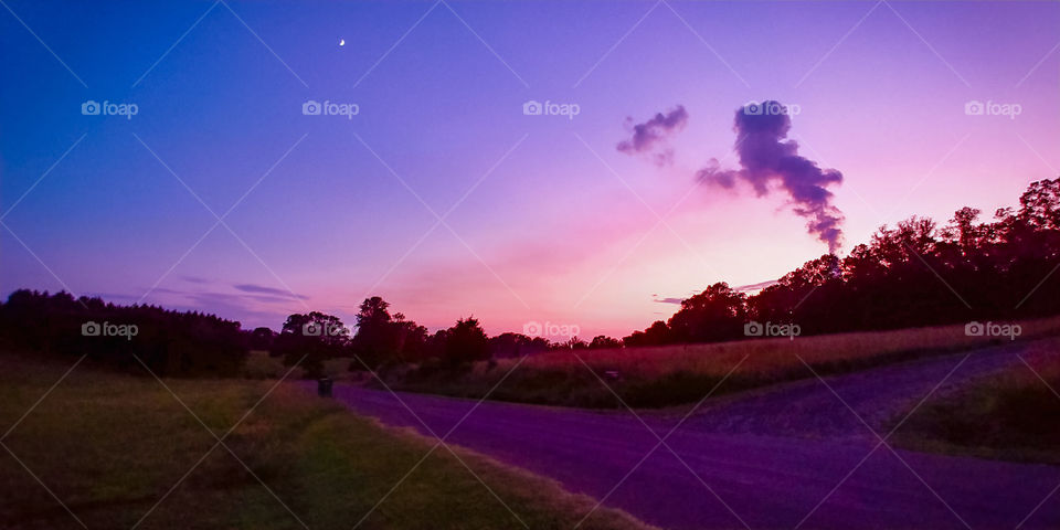 smoke rolling into the evening sky lit up in blues purples and pinks. crescent moon in the distance. country roads and fields of straw.