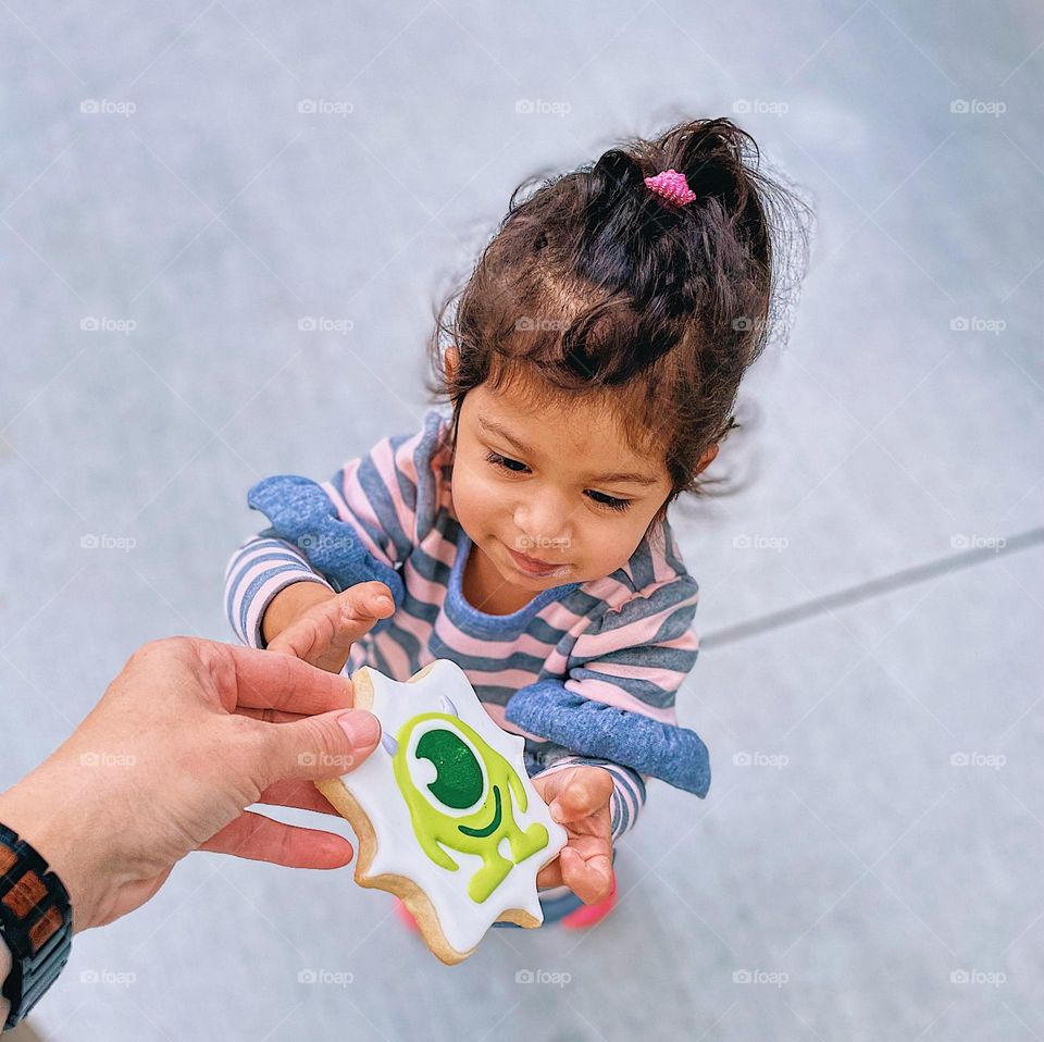 Mother handing daughter a cookie, toddler taking frosted cookie from mother, enjoying Monsters Inc. cookies, toddlers pure joy with cookie, moments of happiness with children 