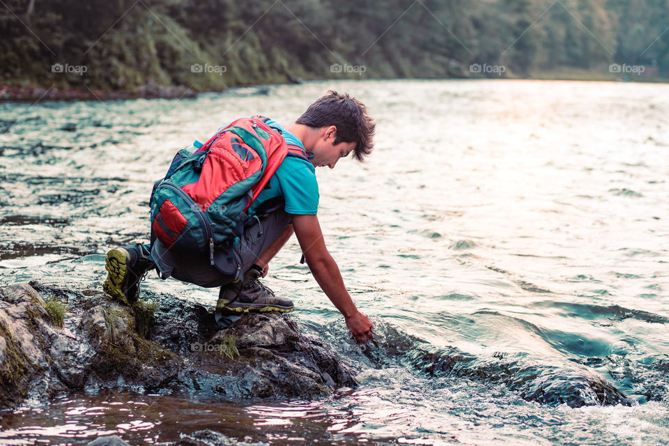 Young boy with backpack sitting on rock over a river, holding hand in water, looking at mountains peaks, he is wearing sports summer clothes